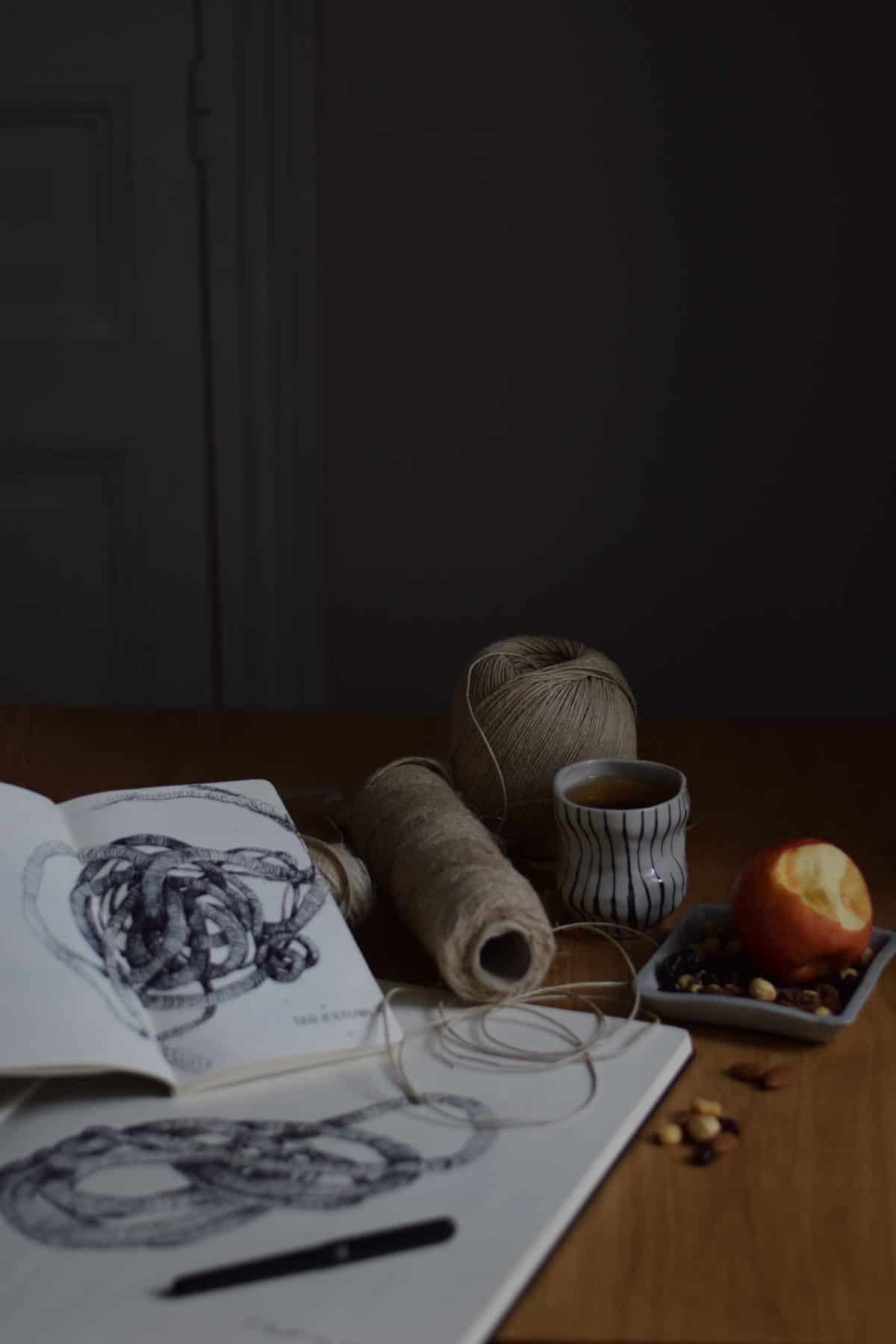 wabi-sabi shot of Aude Franjou's workshop with open notebooks. a half eaten apple and nuts in a bowl, and a few reels of linen string